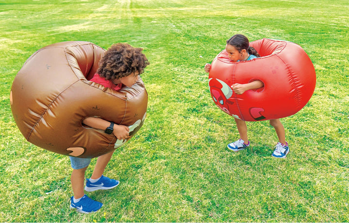 Two children wearing brown and red Rovers blow-up bumper balls playing on green grass outdoors