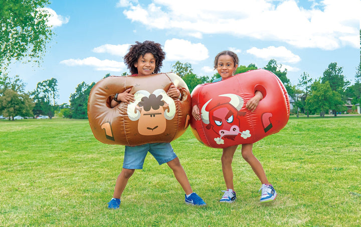 Two children outdoors wearing brown Rovers blow-up bumper balls with cartoon ram and red bull designs on grass